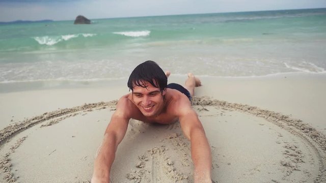 Young happy handsome man lies on the beach draws a heart symbol on the wet sand on the seashore. slow motion. 1920x1080