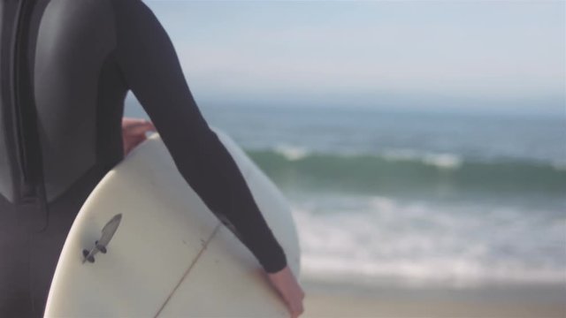 Young man picks up his surfboard, and walks out of focus on a beach shore