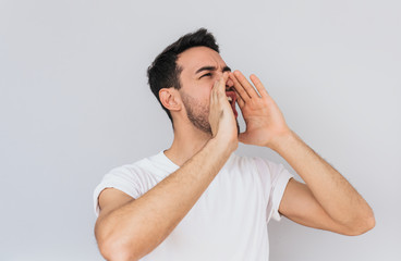 Handsome aggressive male opens mouth widely, screams in panic with hands on mouth, being in stressful situation posing over white studio wall background. People and emotion concept.