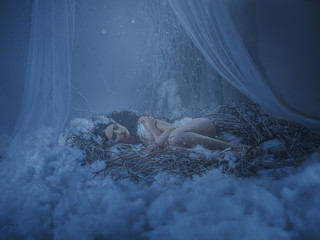 The girl bird sleeps in her nest, she froze, and shrank into the embryo pose in the hope of warming. A beam of light and snow falls on it. Background white wall, twigs, tulle and fluff. Artistic