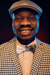 Studio neon portrait of handsome black jazzman with funny smiling face in white shirt and plaid jacket with floral bow tie and vintage cap