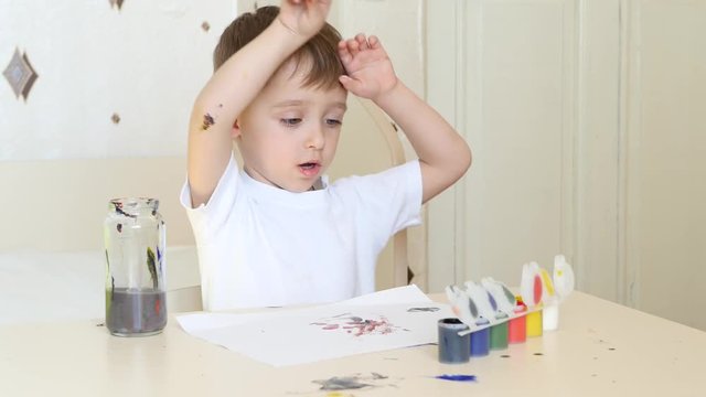 A little boy is sitting at the table and drawing with a brush on a piece of paper. Watercolor paints.