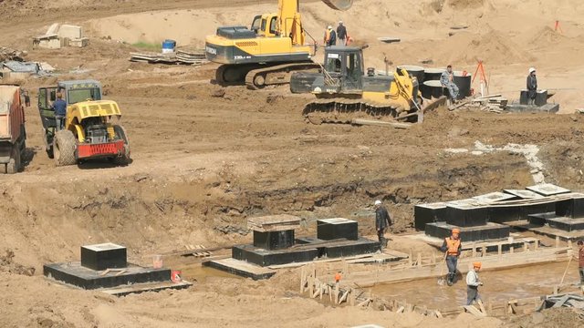 Large construction site. Bulldozer working on construction site. Workers in helmets and suits level surface and pour the foundation for building into wooden formwork outdoors