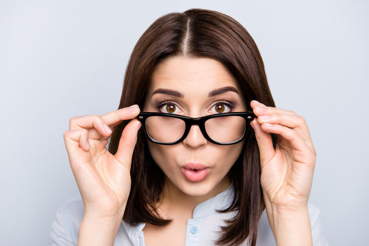 Close up portrait of shocked, impressed, pretty, charming, cute, successful, professional girl looking out glasses, holding eyelets of spectacles on face with two hands, isolated on grey background