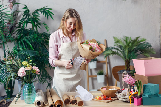 Photo of blonde florist in apron with bouquet with kraft paper on table with paper, marmalade