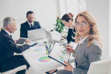 Fototapeta premium Portrait of smart, concentrated, charming, stylish woman sitting with partners together at desktop in workplace, workstation looking at camera having clipboard with diagrams in hands