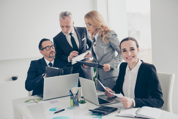 Portrait of pretty, brunette, confident, smiling woman in suit sitting at desktop with colleagues who discuss project, holding paper in hands, looking at camera, having laptop on the table