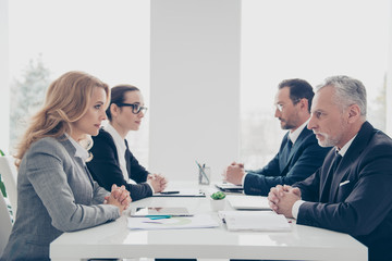 Business competition, four stylish attractive business persons in suits having disagreement and conflict, sitting together at desktop in front of each other, face to face with disrespect expression - obrazy, fototapety, plakaty