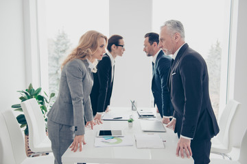 Business competition, four stylish business persons in suits having disagreement and conflict, standing together near desktop in front of each other, face to face with disrespect expression - obrazy, fototapety, plakaty
