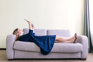 Woman in blue dress lying on the gray couch reading a glossy magazine