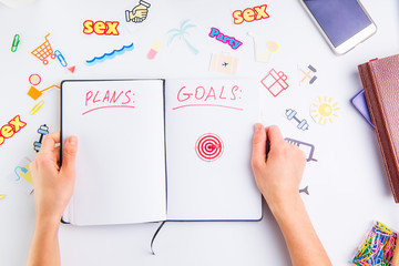 Female hands holding personal organizer with Plans and Goals headline and pushpin as arrow on target icon among other icons of actions on the working place. Concept of planning and goals achievements.