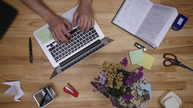 Businessman working on a laptop at office desk with paperwork and other objects around, top view