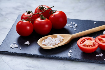 Fresh tomatoes and a spoon of salt on black stony board over white background, close-up, selective focus.