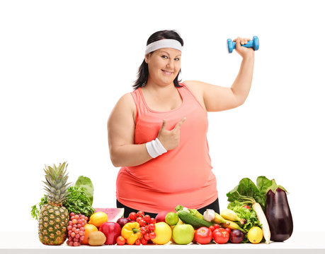 Overweight woman holding a small dumbbell and pointing behind a table with fruit and vegetables