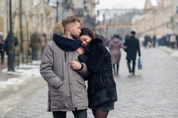 young happy couple hug each other while walking in the street of town in winter under snowfall and smiling, man kiss her girlfriend in temple, girl smile in response