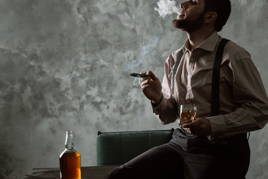 Stylish bearded man sits on the table and drinking whiskey, smoking cigar