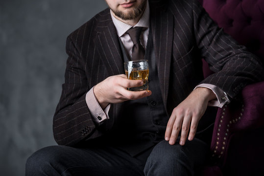 Man in expensive suit is sitting on the leather armchair and drinking whiskey. No face