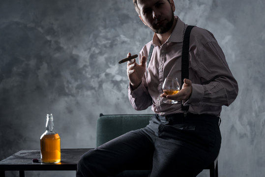 Stylish bearded man sits on the table and drinking whiskey, smoking cigar