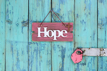 Pink wood sign with the word Hope and a religious cross next to heart lock hanging on rustic antique teal blue wooden door; Easter and Memorial Day holiday concept background with copy space
