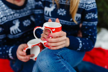 Mug of hot tea with a picture of a heart in the hands of a young couple in warm sweaters sitting on a red blanket. Wedding trip picnic outdoors in the winter in the woods or mountains.