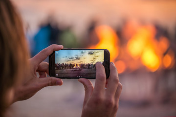 Woman is taking picture of sunset with smart phone. Romantic bonfire night at seaside. People...