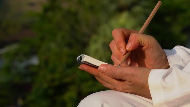 Writing note pad outdoor.
Woman hands holding a small note writing with pencil while sitting on a chair in backyard at sunrise  with green natural blurred background,HD slow motion zoom in.
