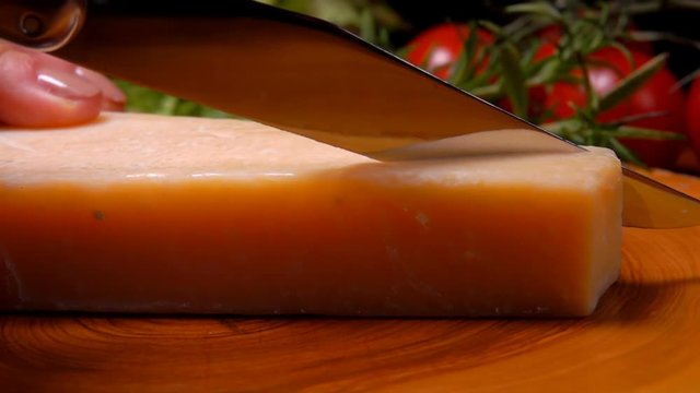 Piece of hard Parmesan cheese cutting by knife on a wooden board on the background of greenery and vegetables