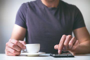 Young man drinking morning coffee and holding mobile phone. Coffee break. Man holding cup of fresh roasted coffee and looking at the smart phone. 
