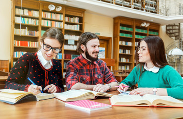 Engaged students checking information in different booksources, two lovely girl wearing cool glasses, bearded boy wearing fashionable checked shirt, indoor shot in college library