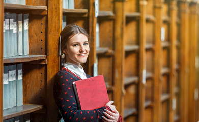 Curious woman standing near library bookshelves with red book