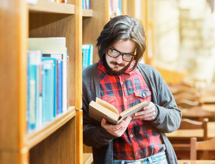 Bearded student man with glasses reads book near bookshelf in the library