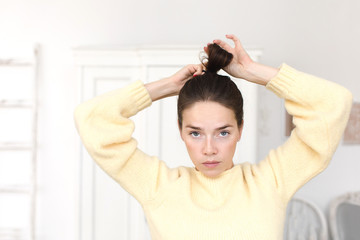 Serious emotionless woman in sweater looking at camera while setting hair in ponytail. 