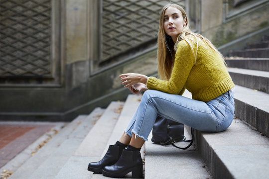 Thoughtful young woman holding smartphone while sitting on steps outdoors