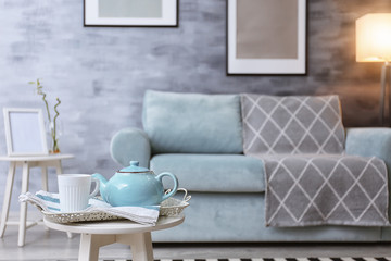 Teapot and cup of hot beverage on small table in room with comfortable sofa