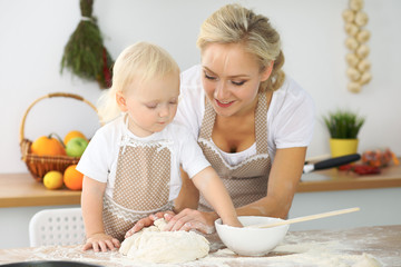 Little girl and her blonde mom in red aprons  playing and laughing while kneading the dough in the kitchen. Homemade pastry for bread, pizza or bake cookies