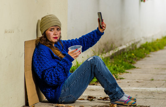 Outdoor view of homeless woman begging on the street in cold autumn weather sitting on the floor at sidewalk taking a selfie