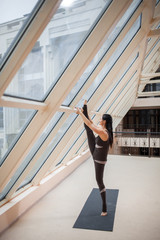 yogi woman in a dark jumpsuit sretching after great workout. Young beautiful young woman in sportswear doing stretching while standing in front of large window