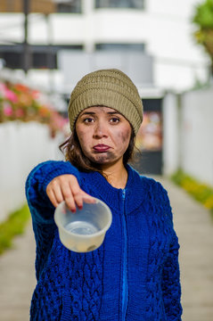 Outdoor view of homeless sad woman on the street in cold autumn weather holding an empty plastic flask in her hands asking for money, at sidewalk