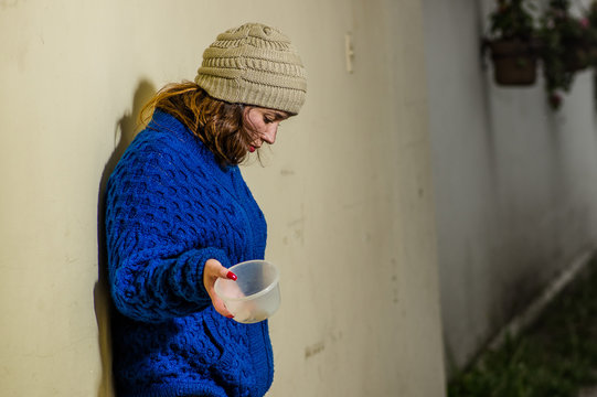 Outdoor view of lonely homeless woman on the street in cold autumn weather holding an empty plastic flask in her hand asking for money, at sidewalk