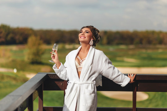 Beautiful young woman in a white robe with a glass of champagne is standing on the terrace and laughing