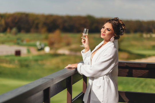 Beautiful young woman in a white robe with a glass of champagne is standing on the terrace and smiling