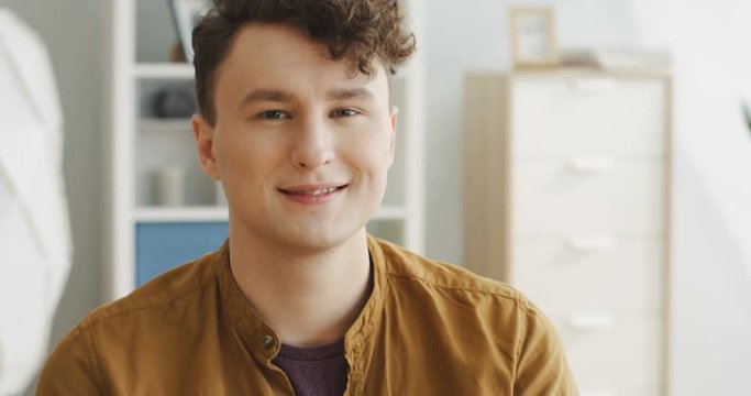 Close up of the young attractive man with a stylish hair looking at the side then turning his head to the camera and smiling in the nice room. Portrait. Inside