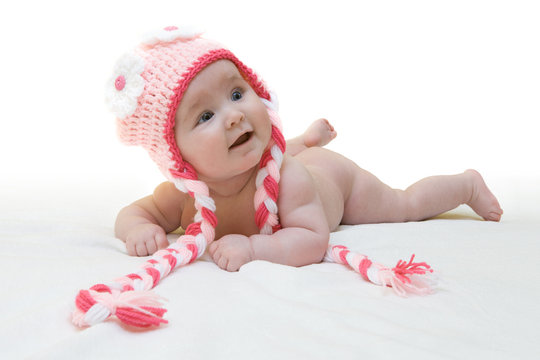 Funny female newborn with nice pink crocheted cap on white background. Happy baby girl with nice crocheted colorful cap on white blanket. Newborn wearing cute crocheted cap with flower ornaments.