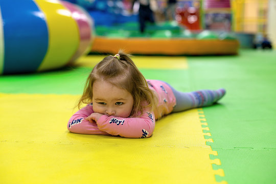 Bored young girl lying on interlocking floor mat in children playgound. Toddler lies on foam mat floor tiles in playroom. Female kid rest on puzzle EVA foam padding. Tired little girl.