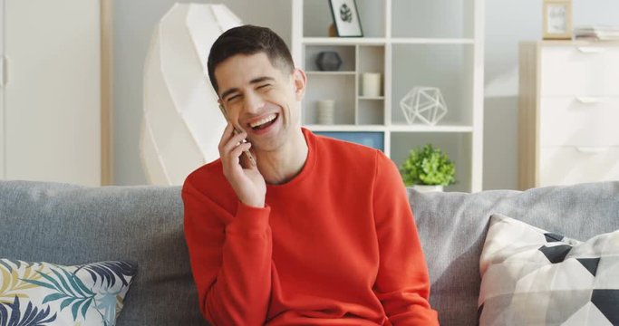 Young handsome man in the red sweater talking on the phone and laughing laud while sitting on the gray sofa in the light living room. Indoor