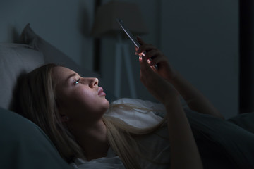 Young sleepy exhausted woman lying in bed using mobile phone, can not sleep. Insomnia, nomophobia,...