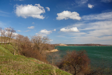 Fototapeta na wymiar Spring landscape by the lake. A bright sunny day, a fresh breeze blows and chases clouds over emerald-colored water. The shore is not yet overgrown with green grass, the trees stand without foliage.