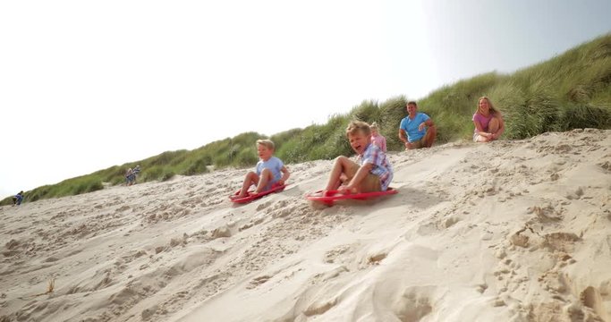 Slow motion shot of two brothers being pushed down a sand dune while sat on sand boards, having a competition.