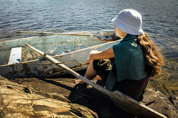 Relax on the shore of a lake. A young woman sits with a paddle on a stone against the backdrop of a vintage wooden boat in the warm light of the day sun.