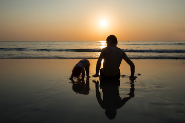 Fototapeta na wymiar Baby girl with her father are sitting on a sand beach and looking to a sea during sunset. Daddy and his little daughter enjoying the sunset at the Andaman Sea.